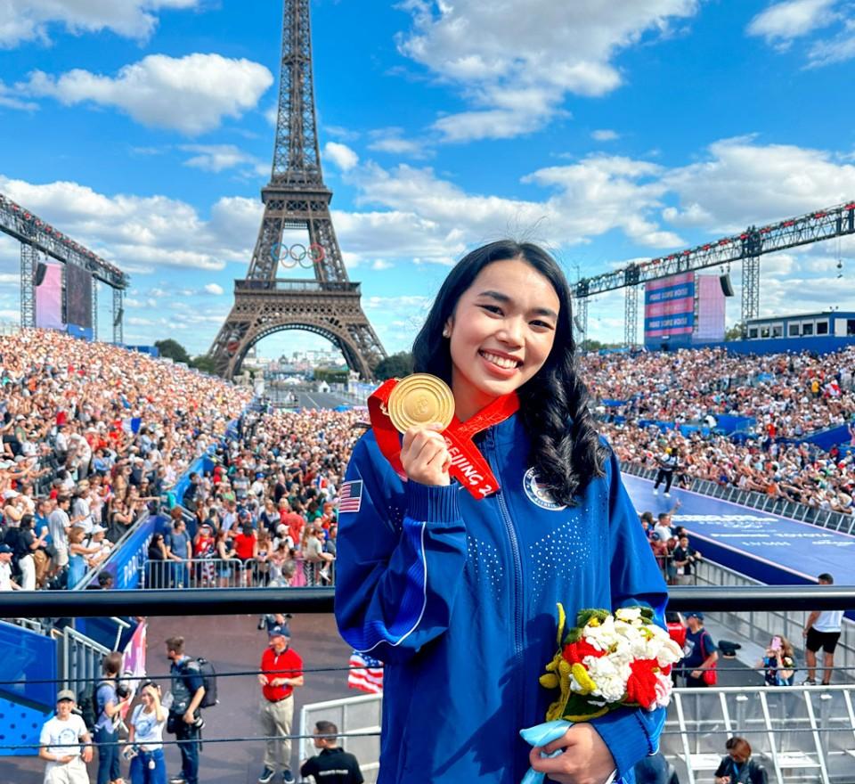 woman holding a gold medal smiling with crowds and the Eiffel Tower behind her