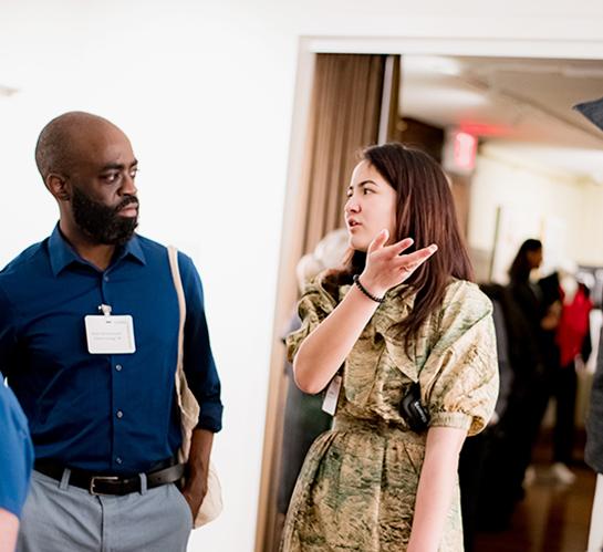 person in a blue shirt speaking to an undergrad student in a print dress next too a mannequin wearing a hooded garment