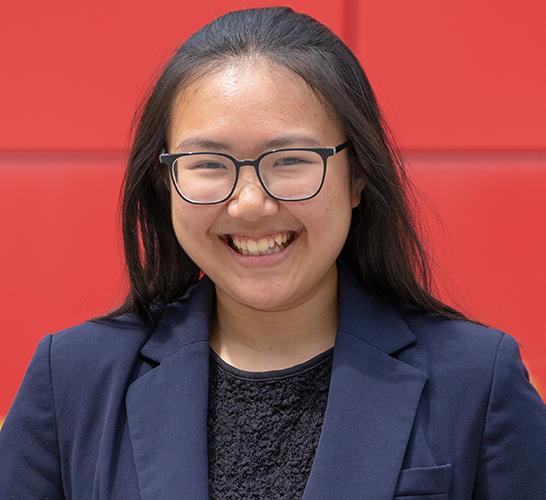 college senior wearing glasses and a blue blazer smiling with a red background