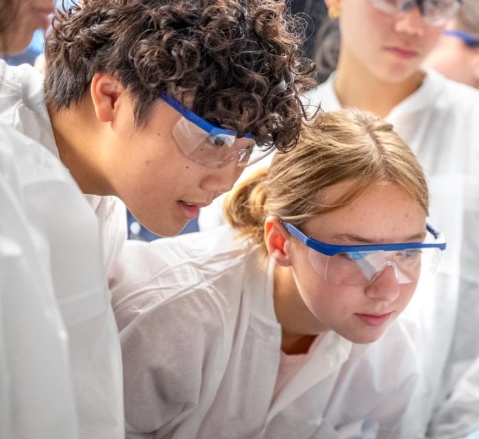 Teens look at an experiment while wearing safety glasses and lab coats