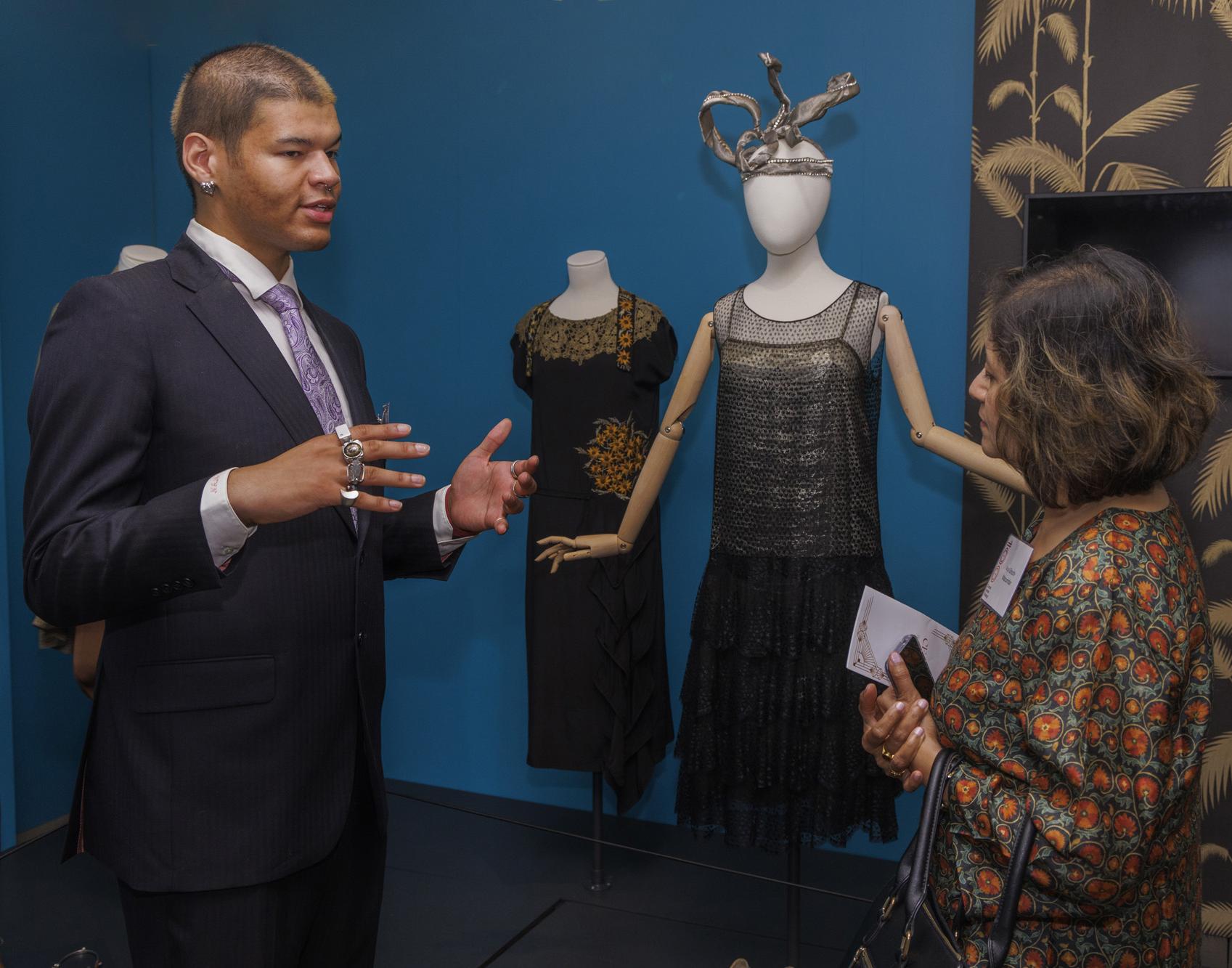 young person in a suit speaks to a woman as they stand in front of mannequins wearing clothing from the 1920s