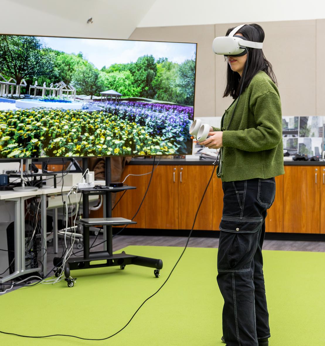 graduate student in a VR headset stands next to a large screen showing a garden