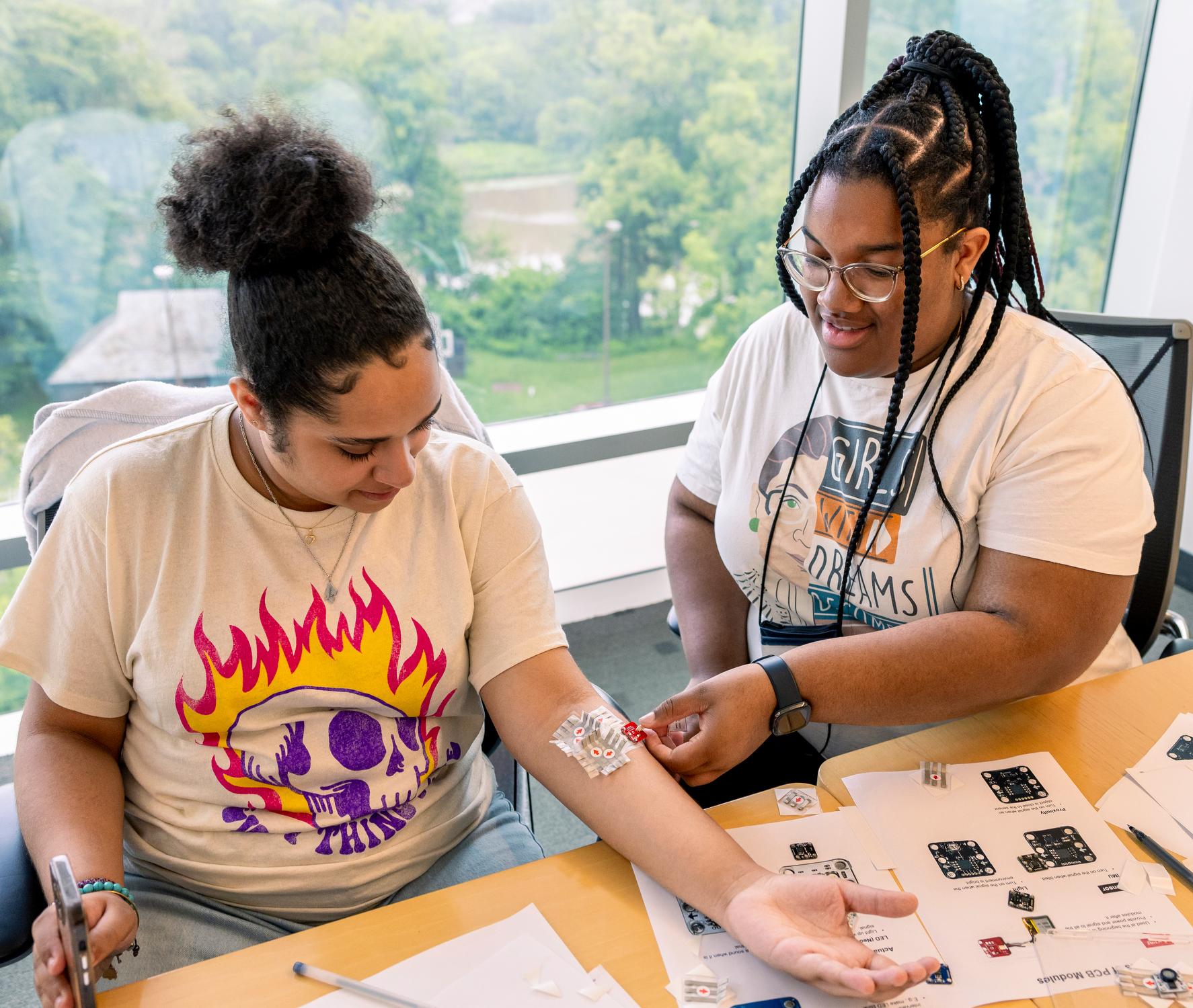 two high school students sitting at a table. one is attaching a sensor to the other's forearm