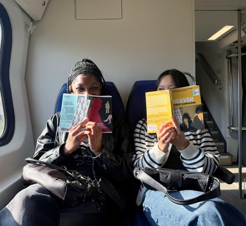 Two women sit on a train in Europe with books in front of their faces 