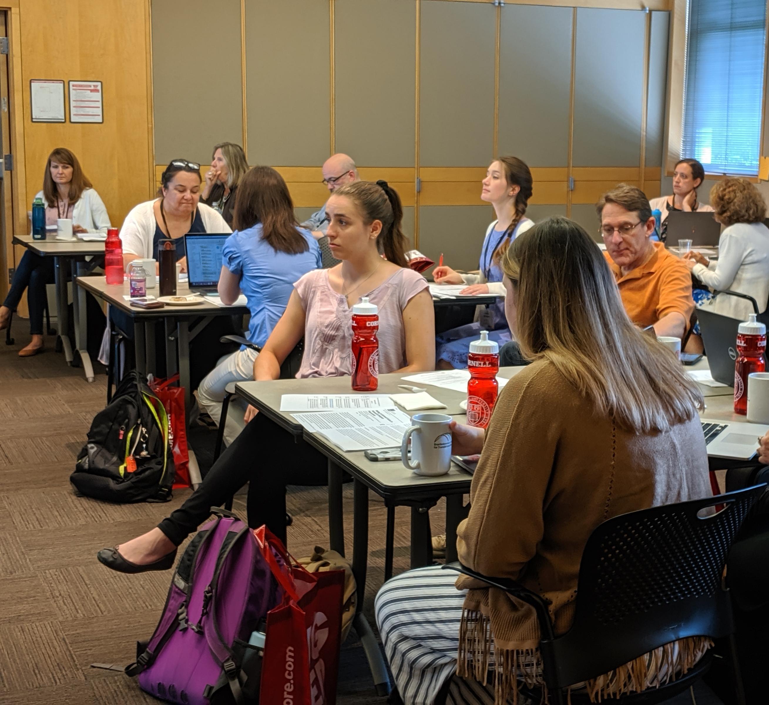 Attendees to a seminar, sitting at tables, listen to a speaker.