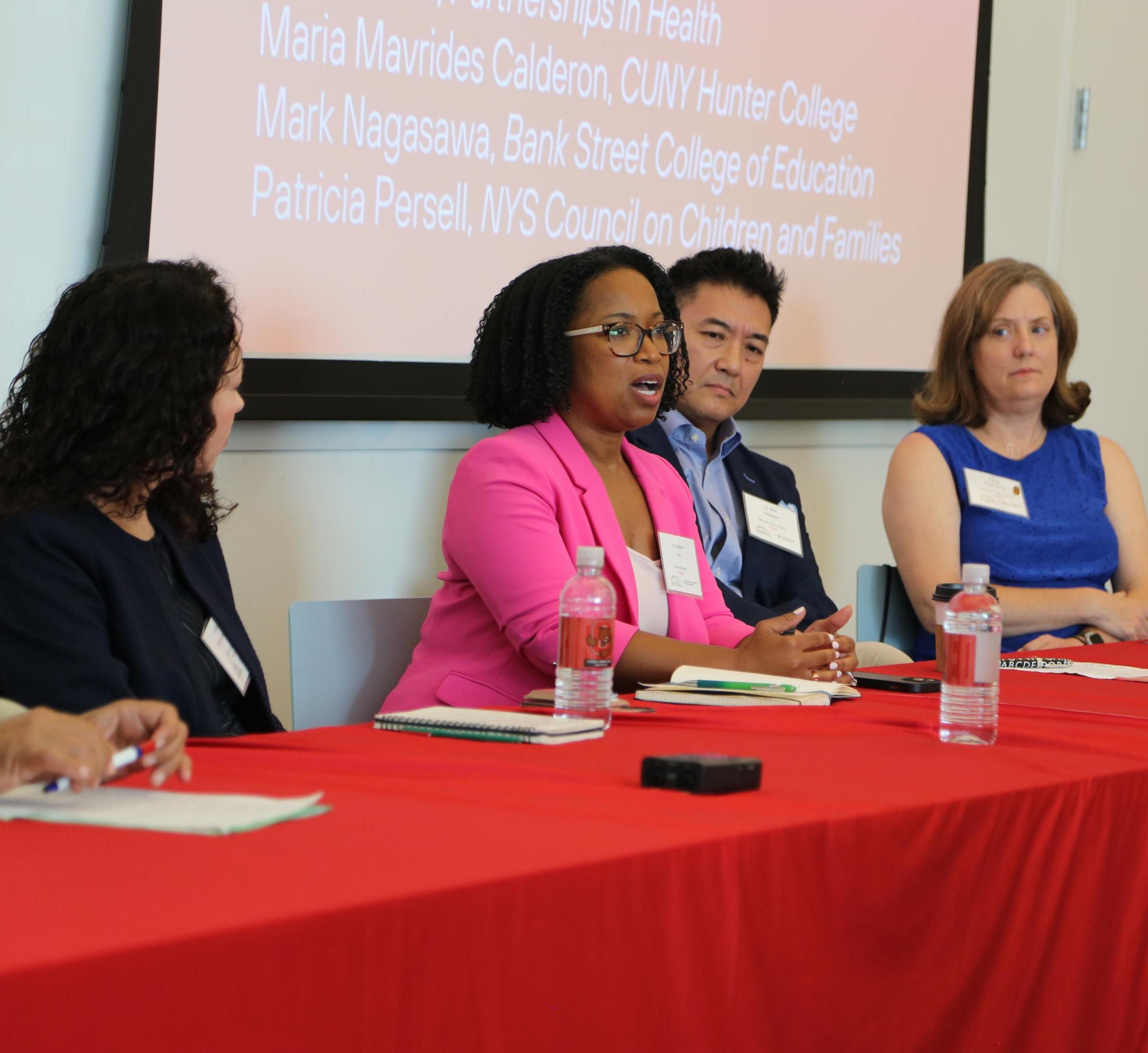A woman in a pink blazer is at a table with three other people. She is giving a lecture.