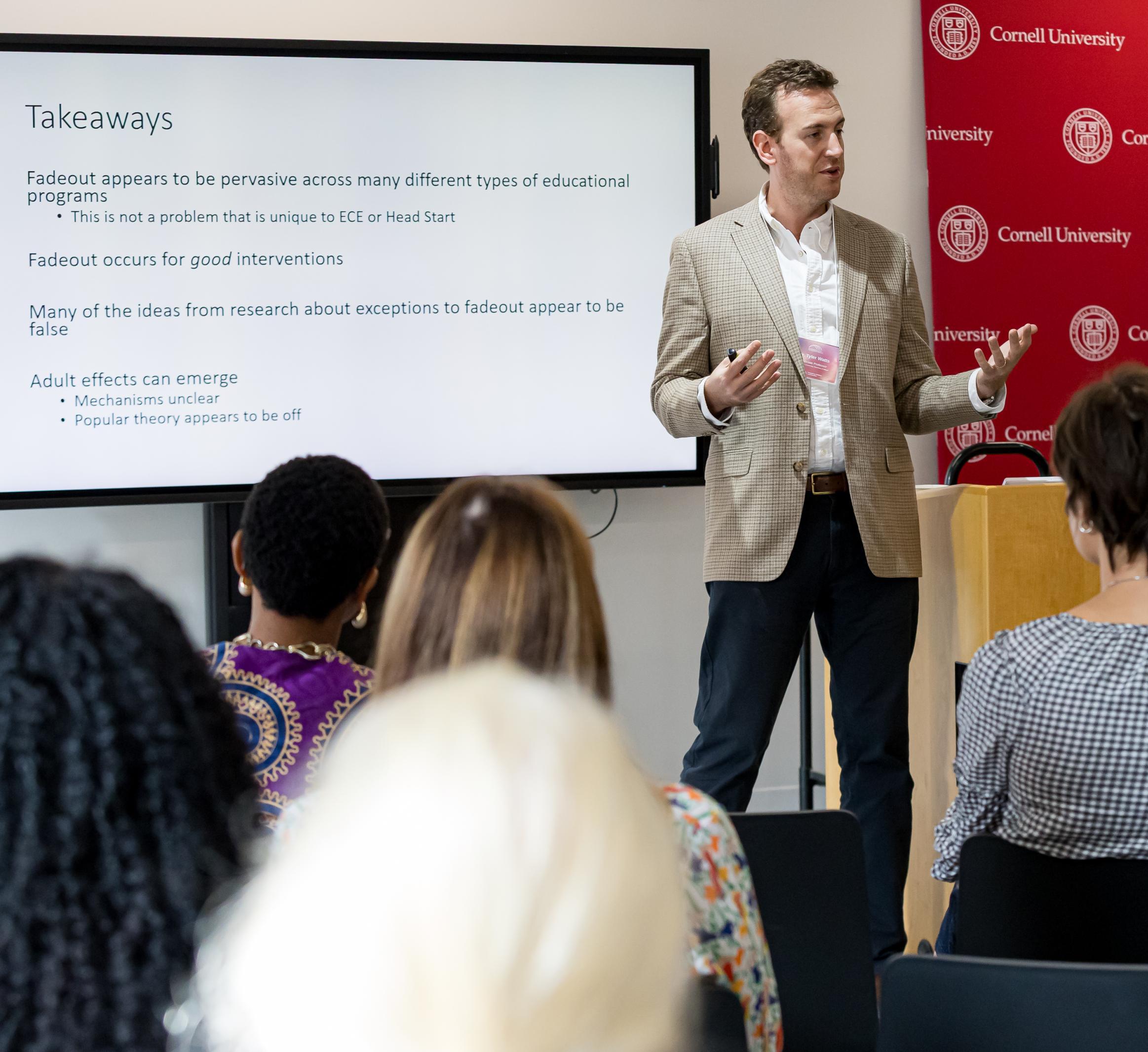 A man stands in a white shirt, tan sportcoat, and black pants, stands in the front of the room giving a lecture to an audience.