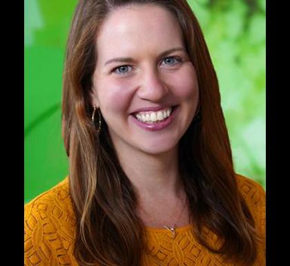 Susan Wiener has long brown hair and is wearing a dark gold sweater. She is standing in front of a green background.