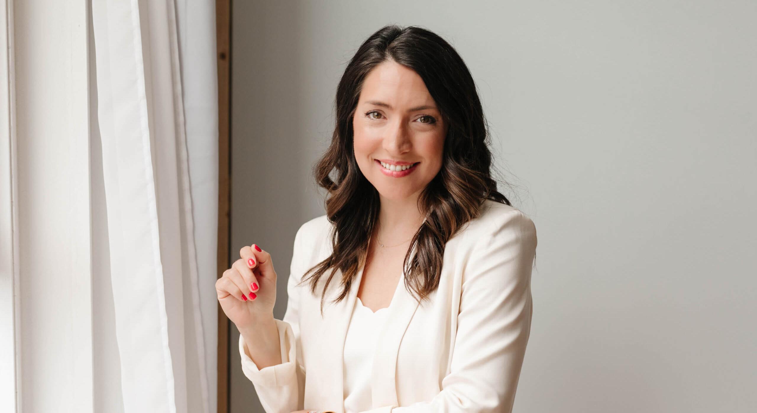 A woman stands in front of a wall with a curtain beside her