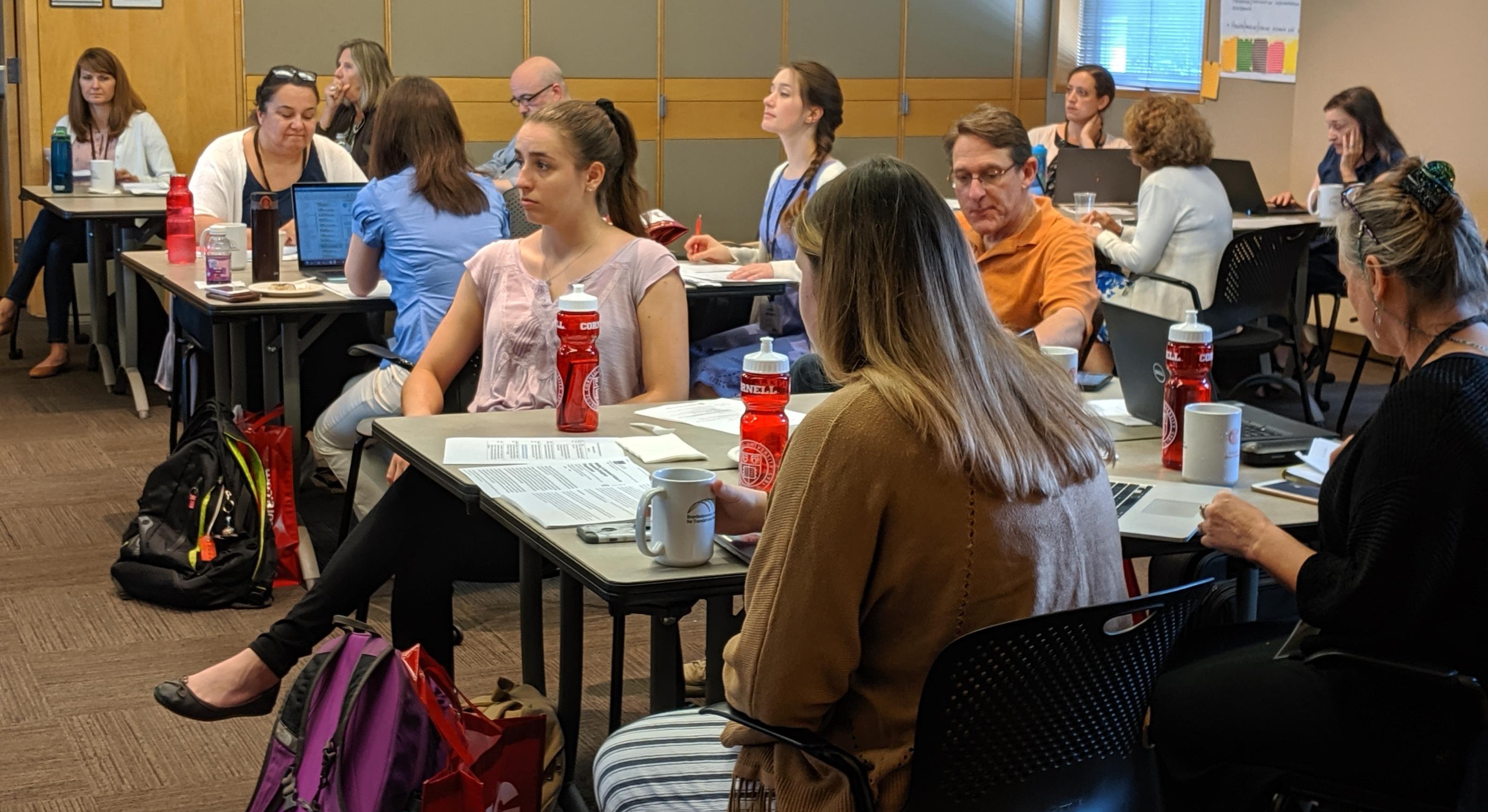 Attendees to a seminar, sitting at tables, listen to a speaker.