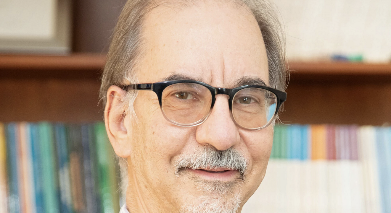 A man with glasses, wearing a grey suit, striped shirt and dark brown tie stands in front of a bookshelf