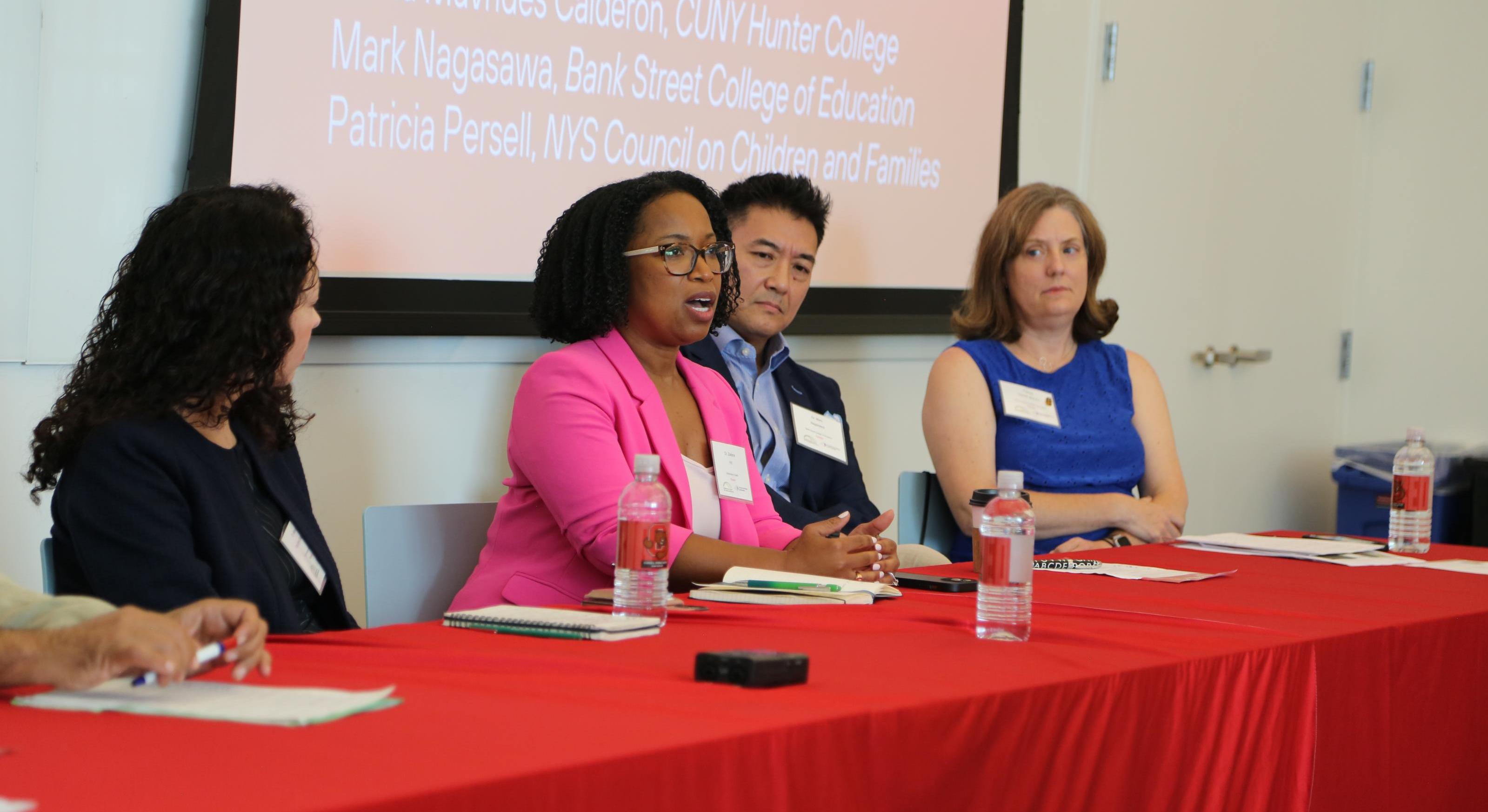 A woman in a pink blazer is at a table with three other people. She is giving a lecture.