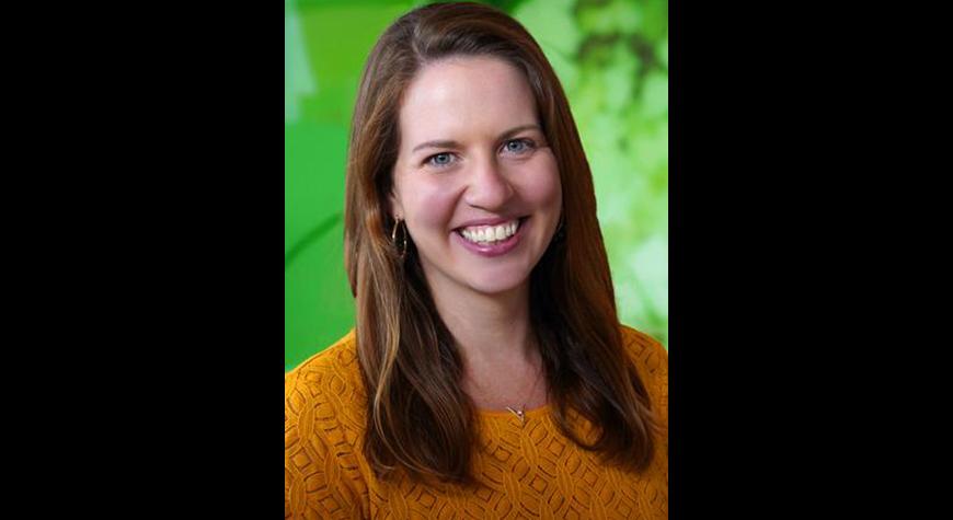 Susan Wiener has long brown hair and is wearing a dark gold sweater. She is standing in front of a green background.