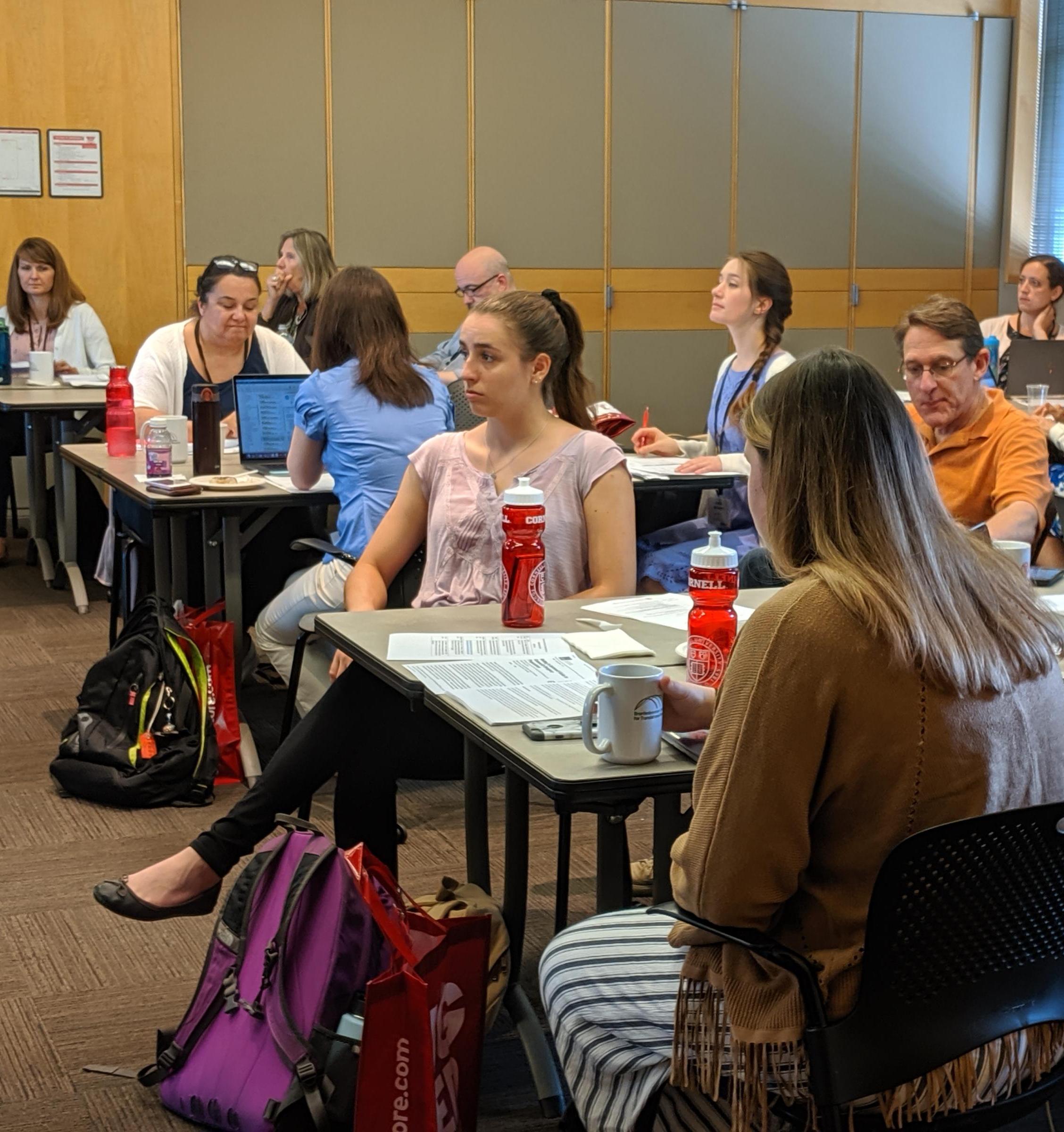 Attendees to a seminar, sitting at tables, listen to a speaker.