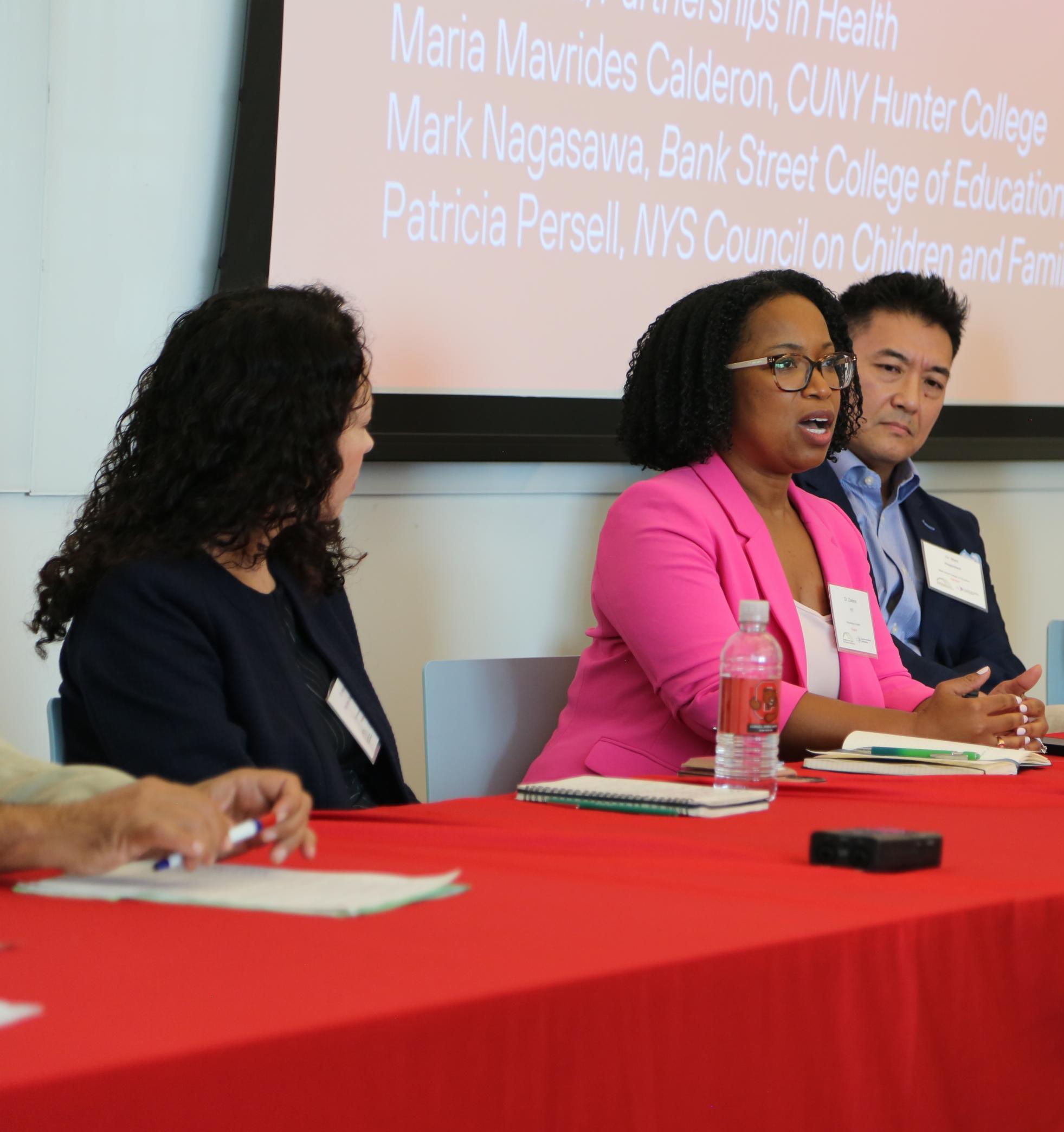 A woman in a pink blazer is at a table with three other people. She is giving a lecture.