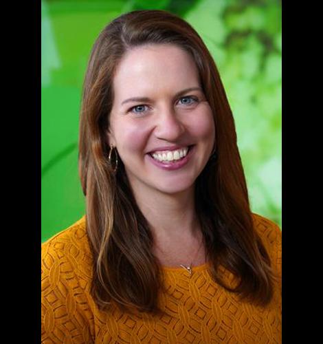 Susan Wiener has long brown hair and is wearing a dark gold sweater. She is standing in front of a green background.