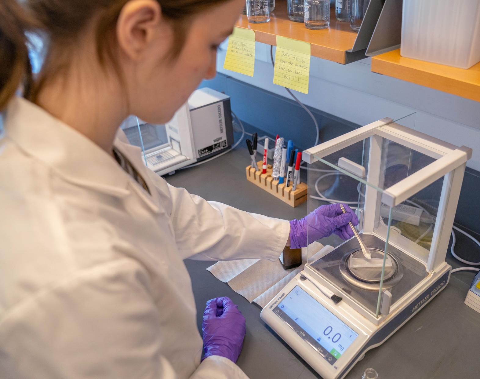 college student in a lab coat adds material to a scale