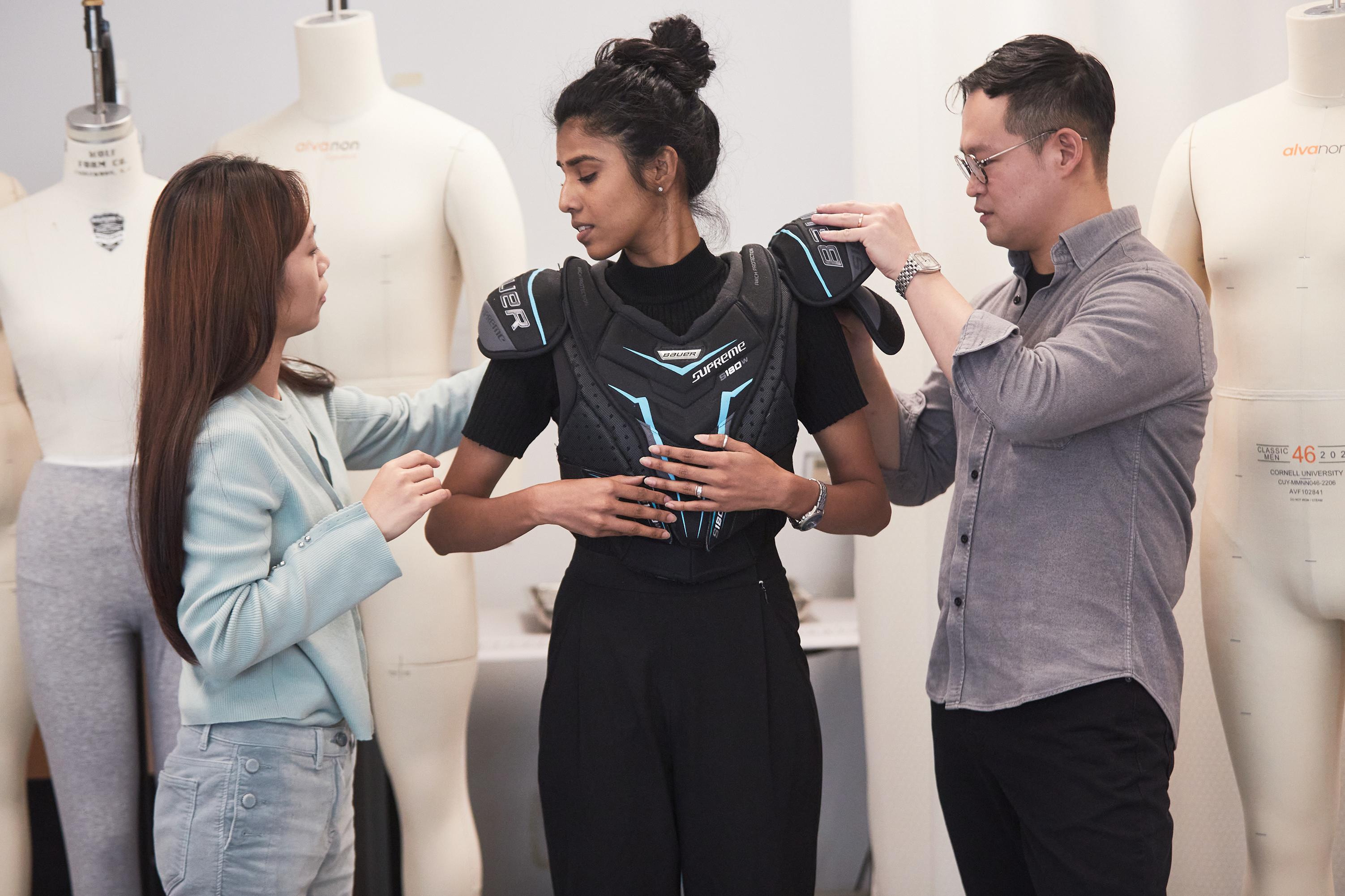 two people adjust protective padded gear to the upper body of a third person. They all stand in front of a row of mannequins