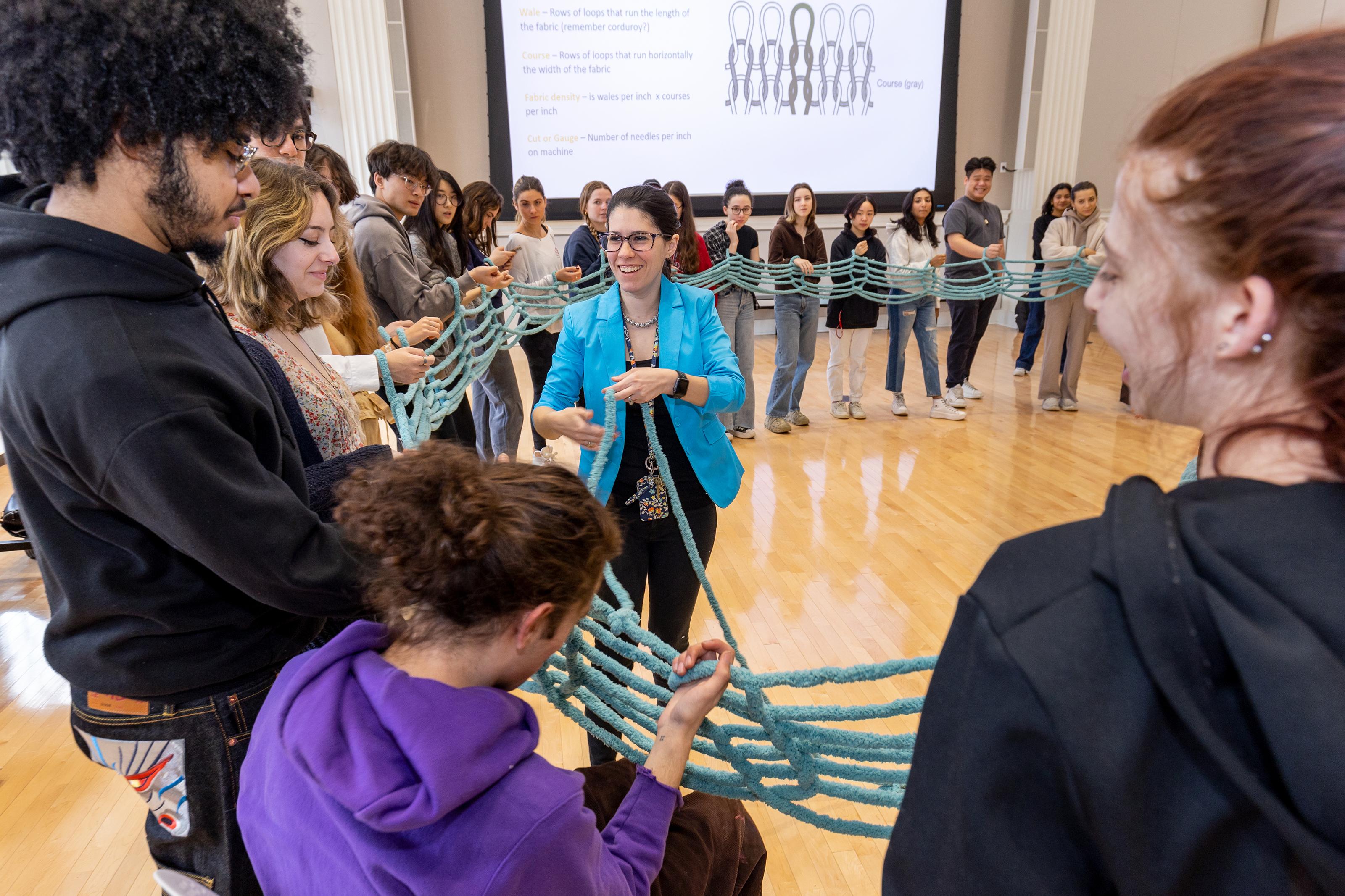 large circle of students stand holding lengths of thick blue yarn which they are knitting together