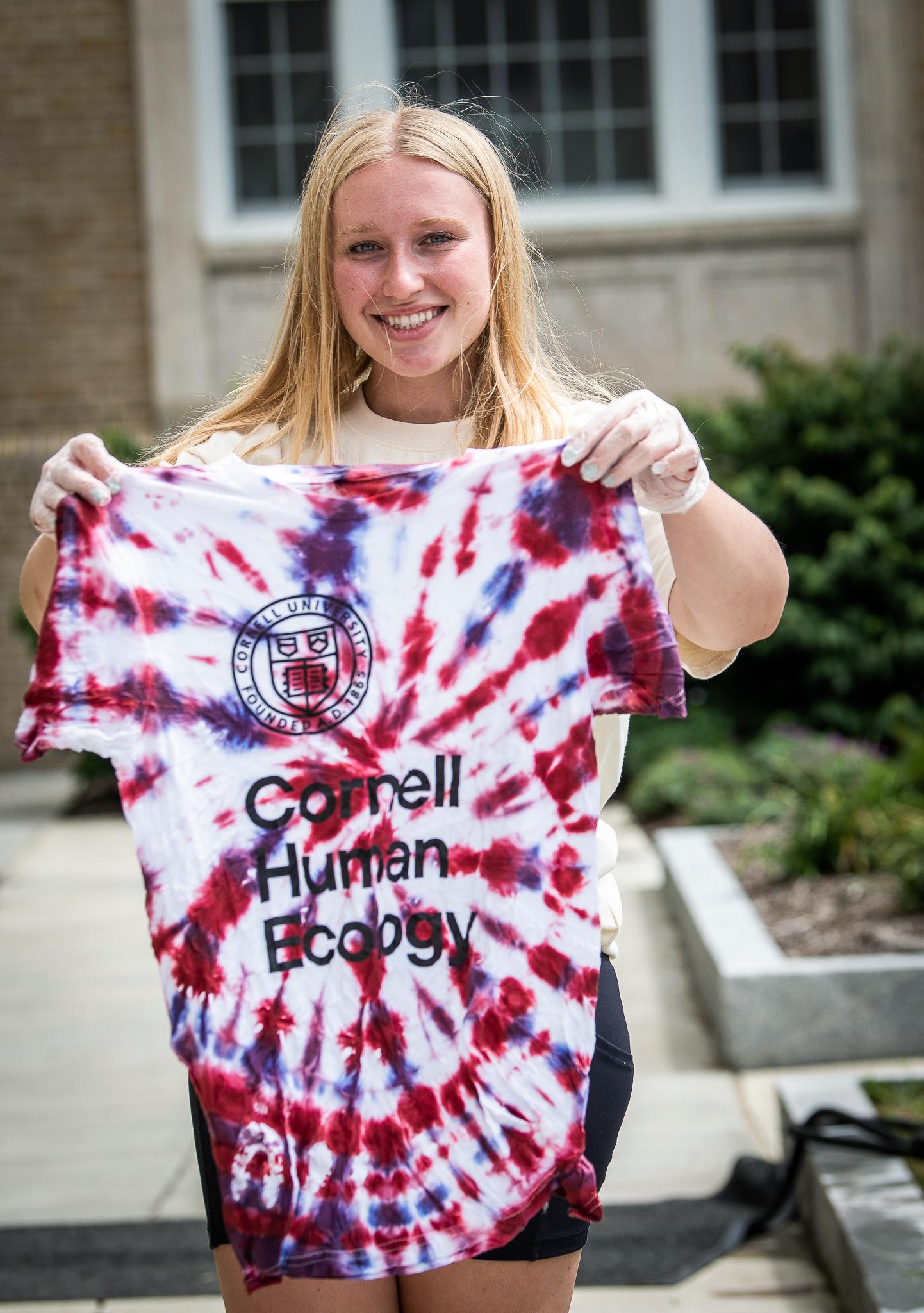 student holds up a white t-shirt with purple and red tie-dyed swirls and a black logo reading "Cornell Human Ecology"