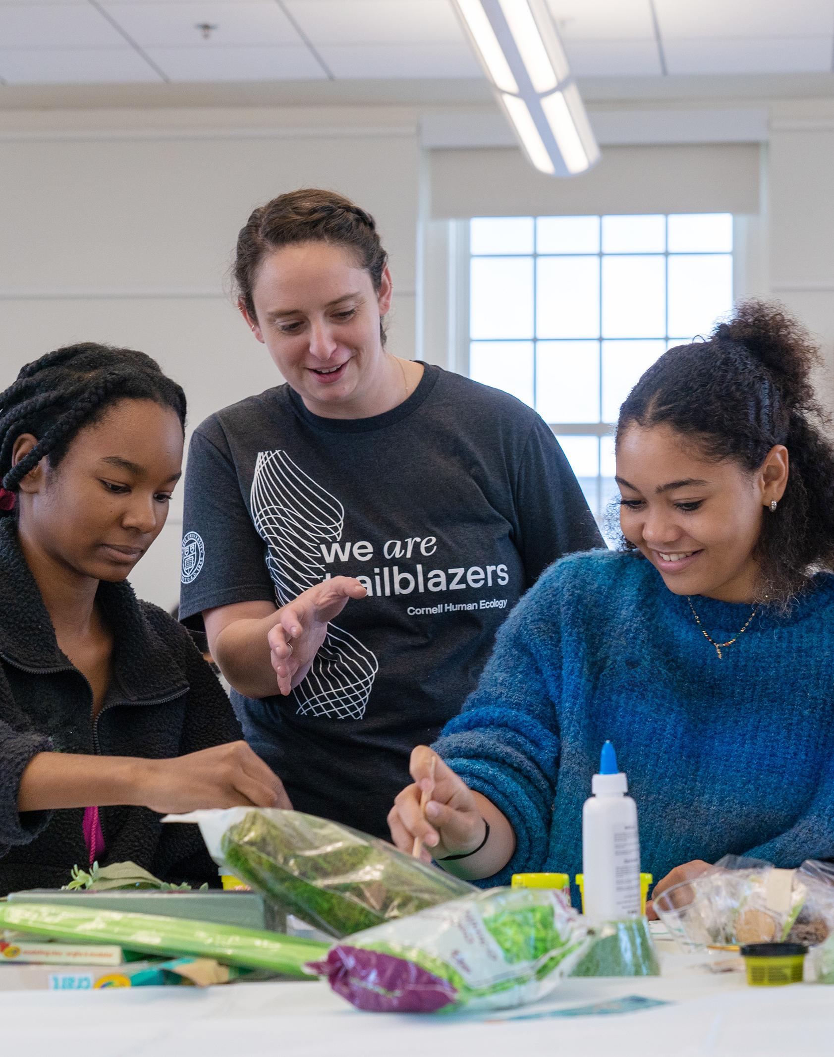 high school students work on an art project at a table while college students stand and chat with them