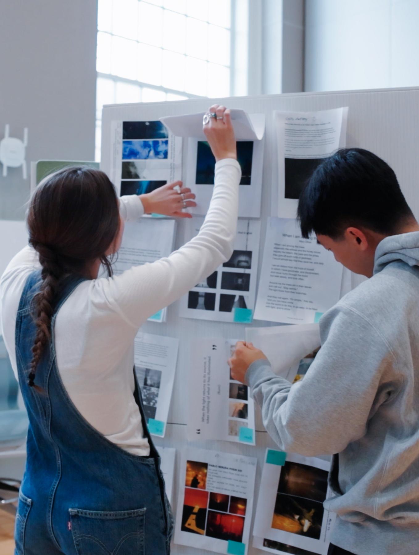 two students pinning papers to a board in a big, bright room
