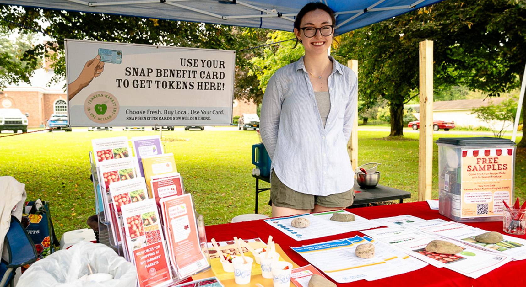 Student Casey Martin stands at a tent teaching people about using SNAP at farmer's market and offering fresh tastings.