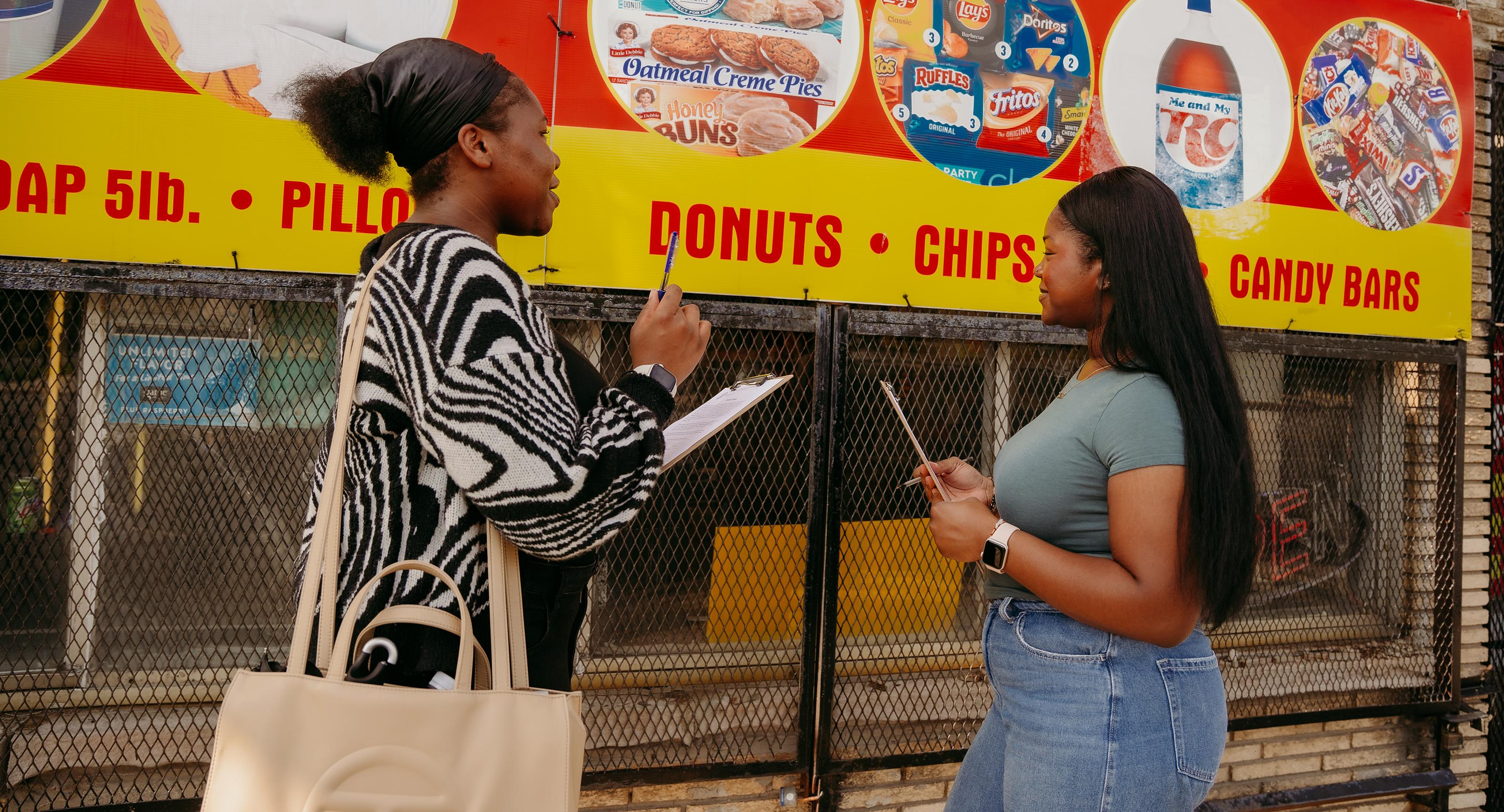 two college students stand on a city street with a colorful sign displaying pictures of snack food behind them