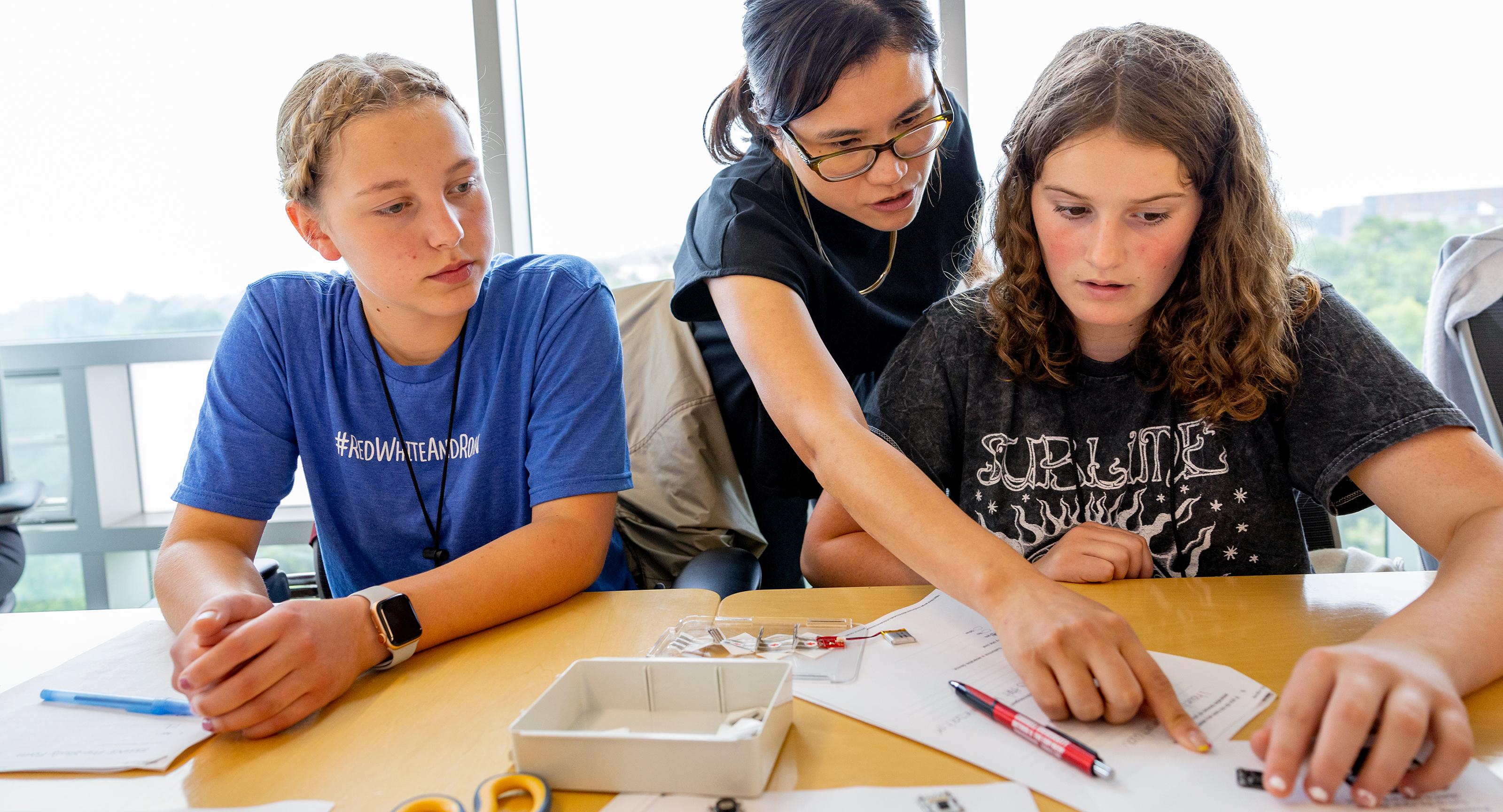 professor leaning over two middle schoolers pointing at a paper on a desk