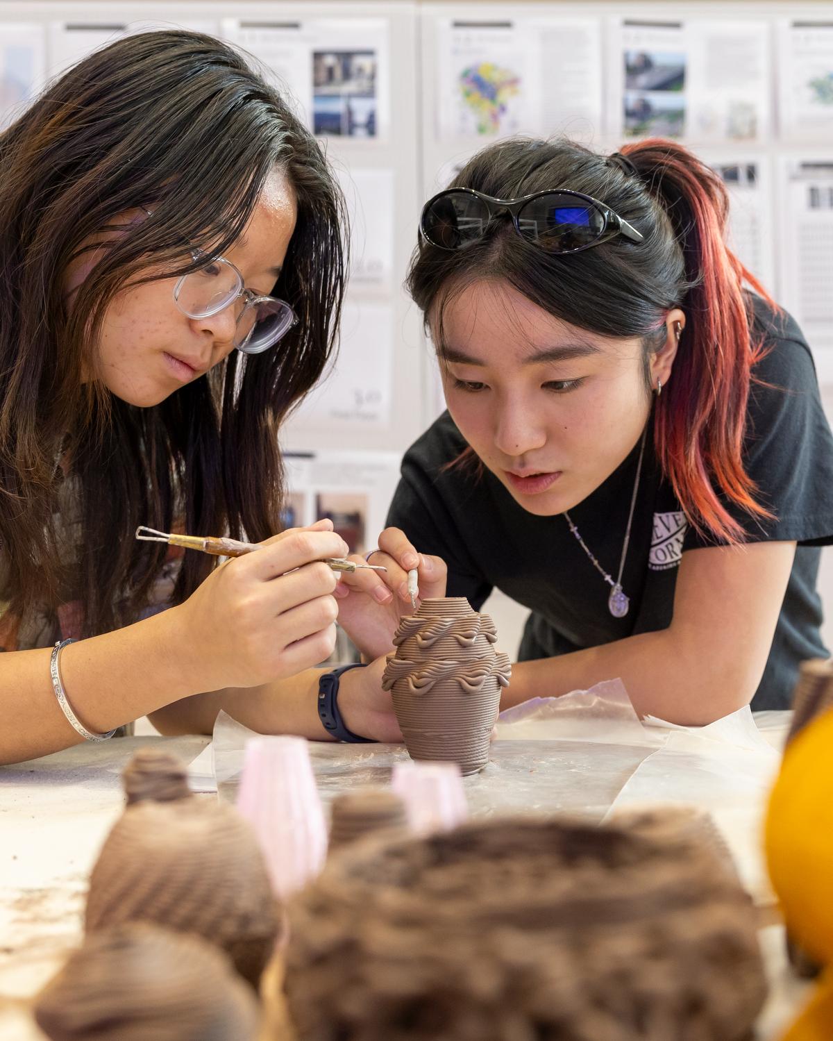 two college students use a stylus to shape a clay object