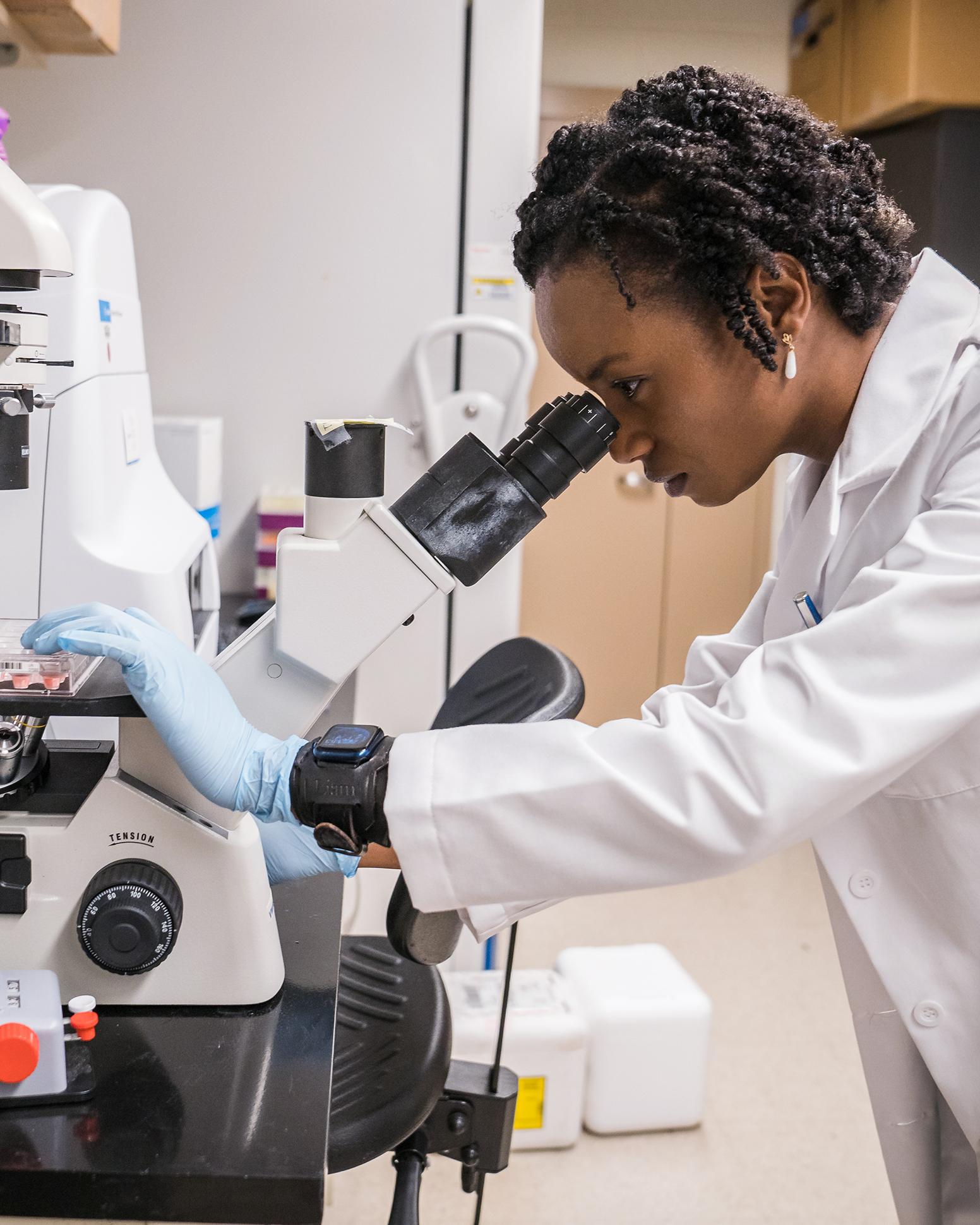 woman in a lab coat leans to look into a microscope in a lab