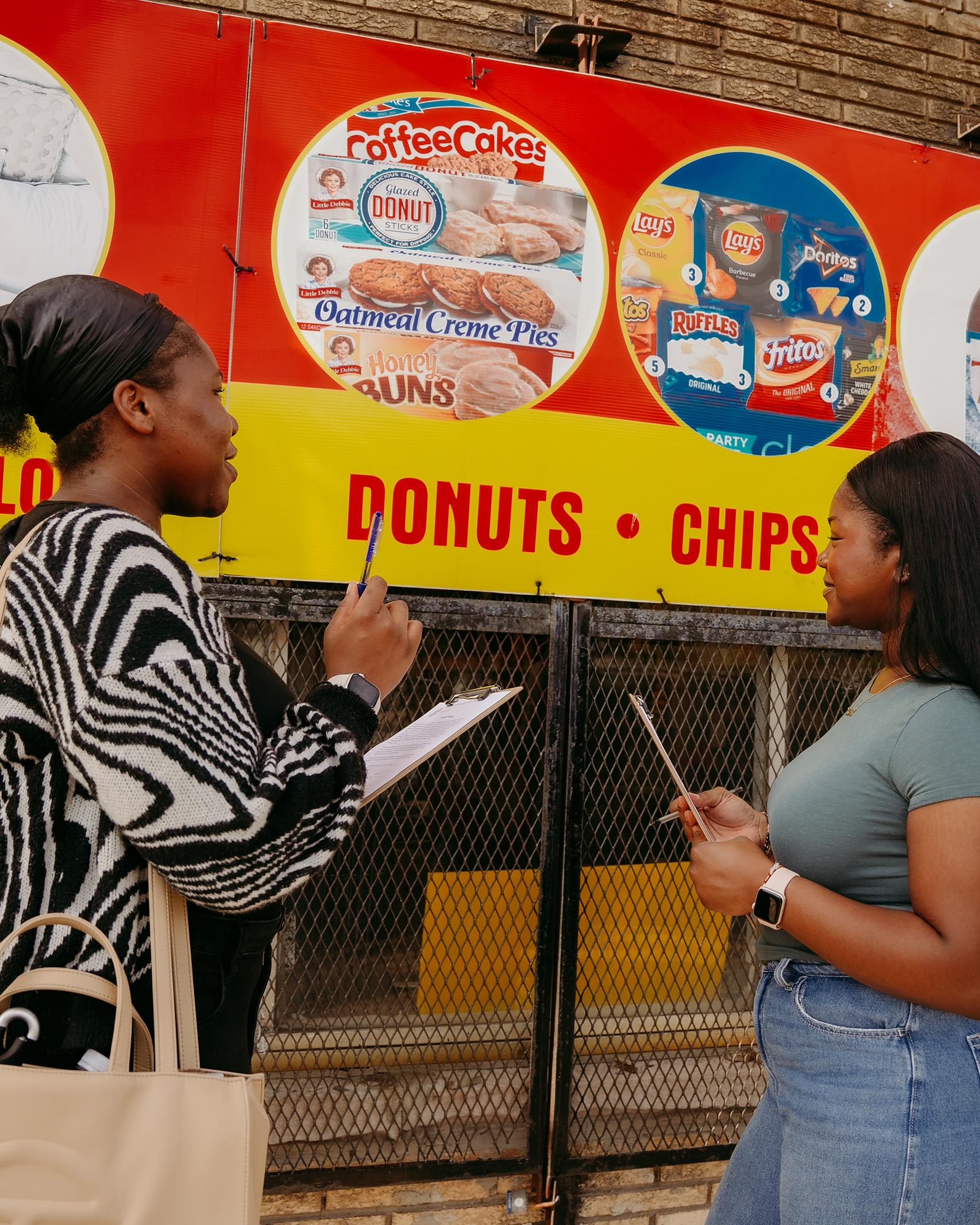 two college students with clipboards talk in front of a sign showing snack foods on a city street