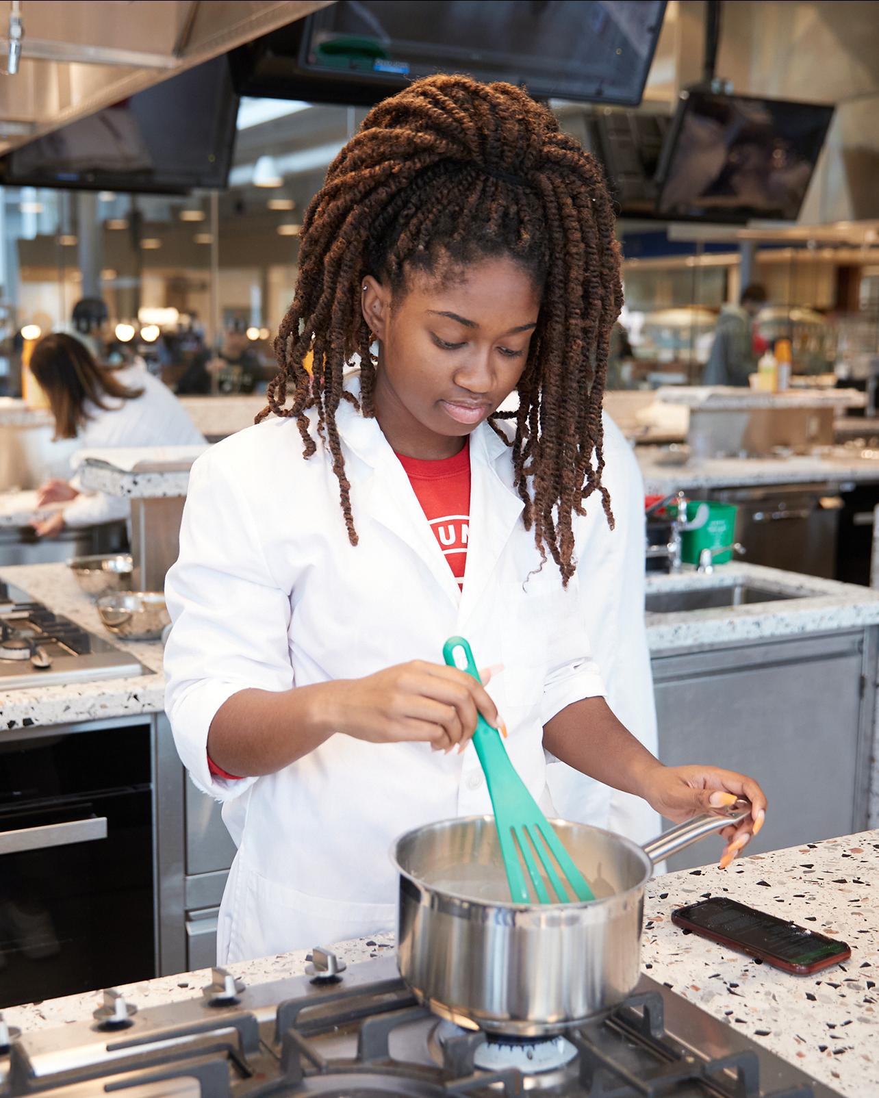 student in a white lab coat working in a modern large teaching kitchen