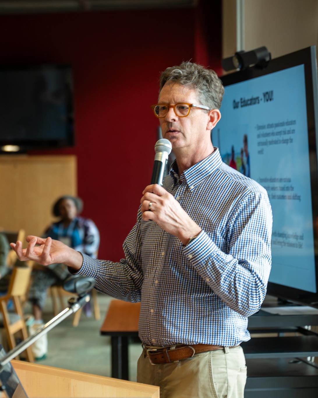 A man with glasses, wearing a blue and white checkered shirt, speaks to an audience in a room