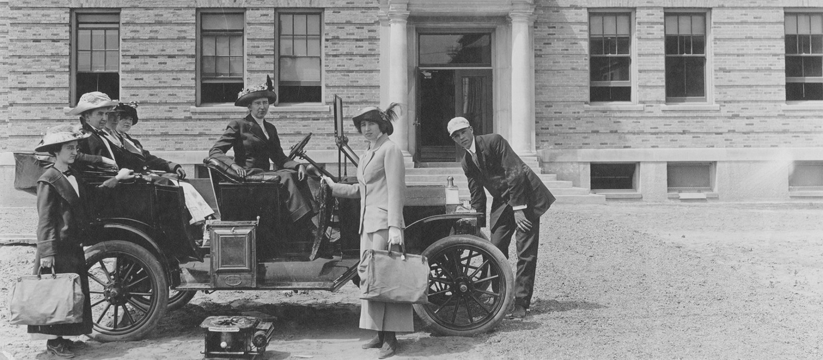 Women and a man in the 1920s around an open car in front of a brick building