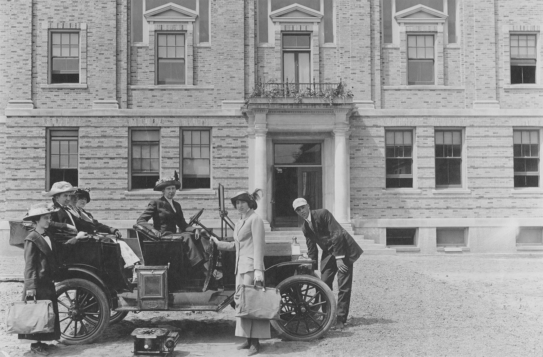 A group of women and a man prepare to drive in an open car in front of a brick building
