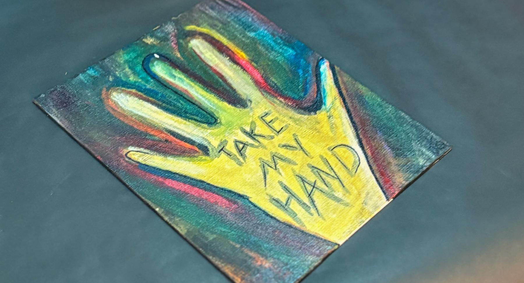 An art piece that is an outline of a hand, colored in yellow, with the words "Take my hand" written inside the hand.