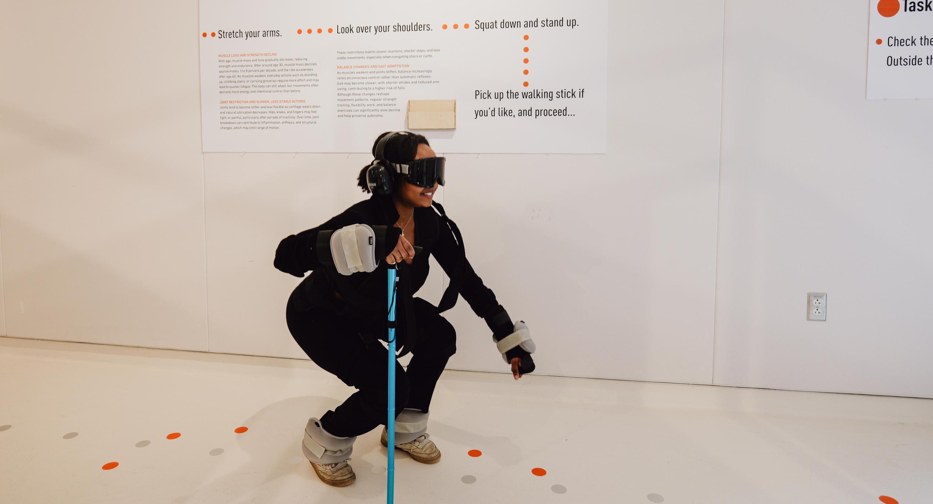 A participant bending down in a simulation suit and holding a walking stick