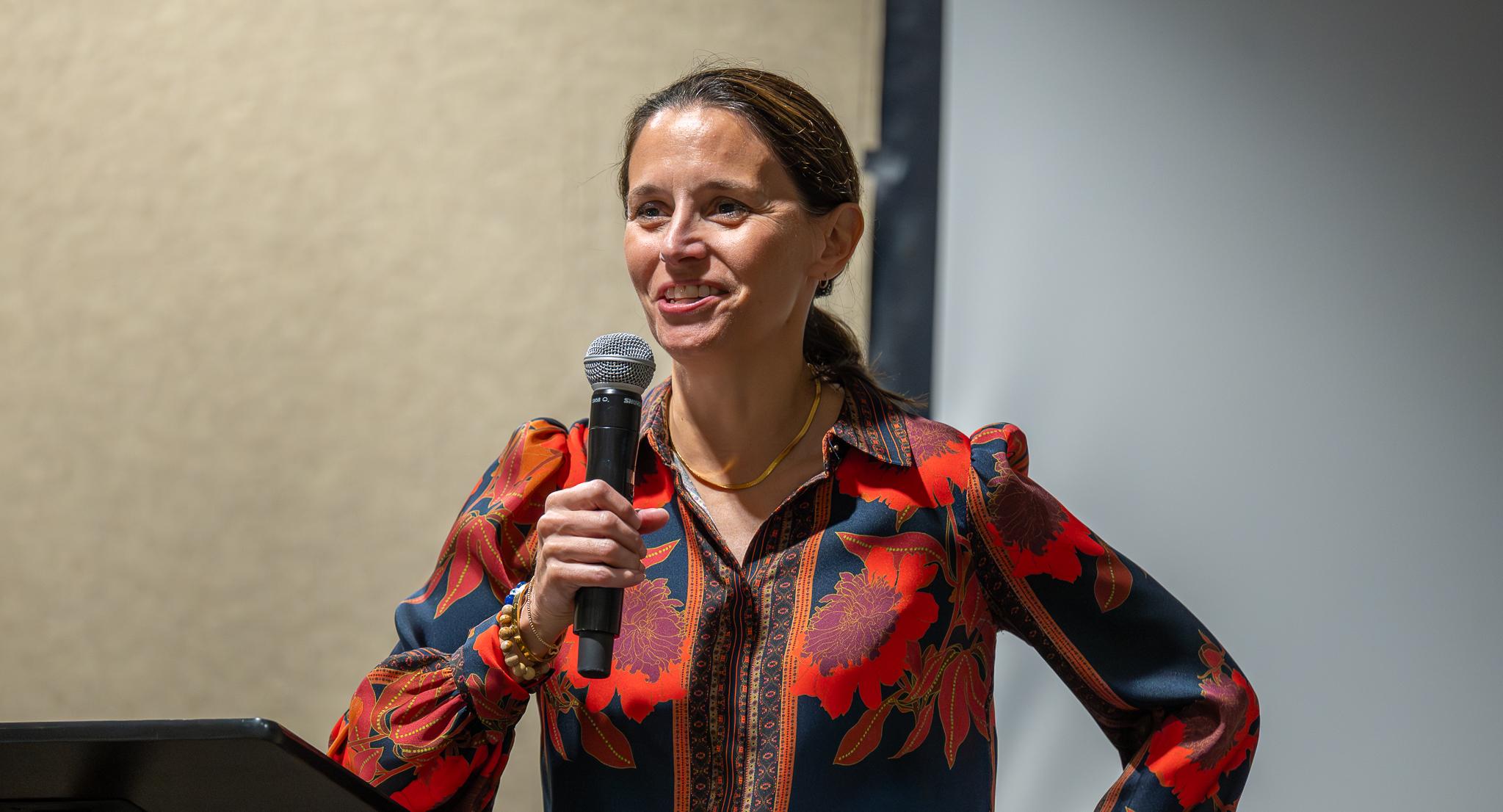 A woman in a red and black shirt, holding a microphone, gives remarks.