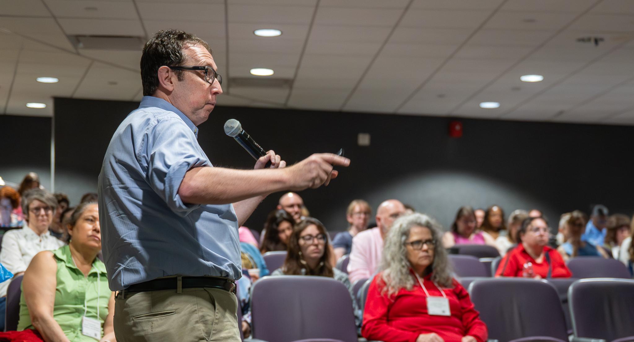 A person wearing a blue shirt and beige pants, with glasses, gives a presentation with an audience in front of him