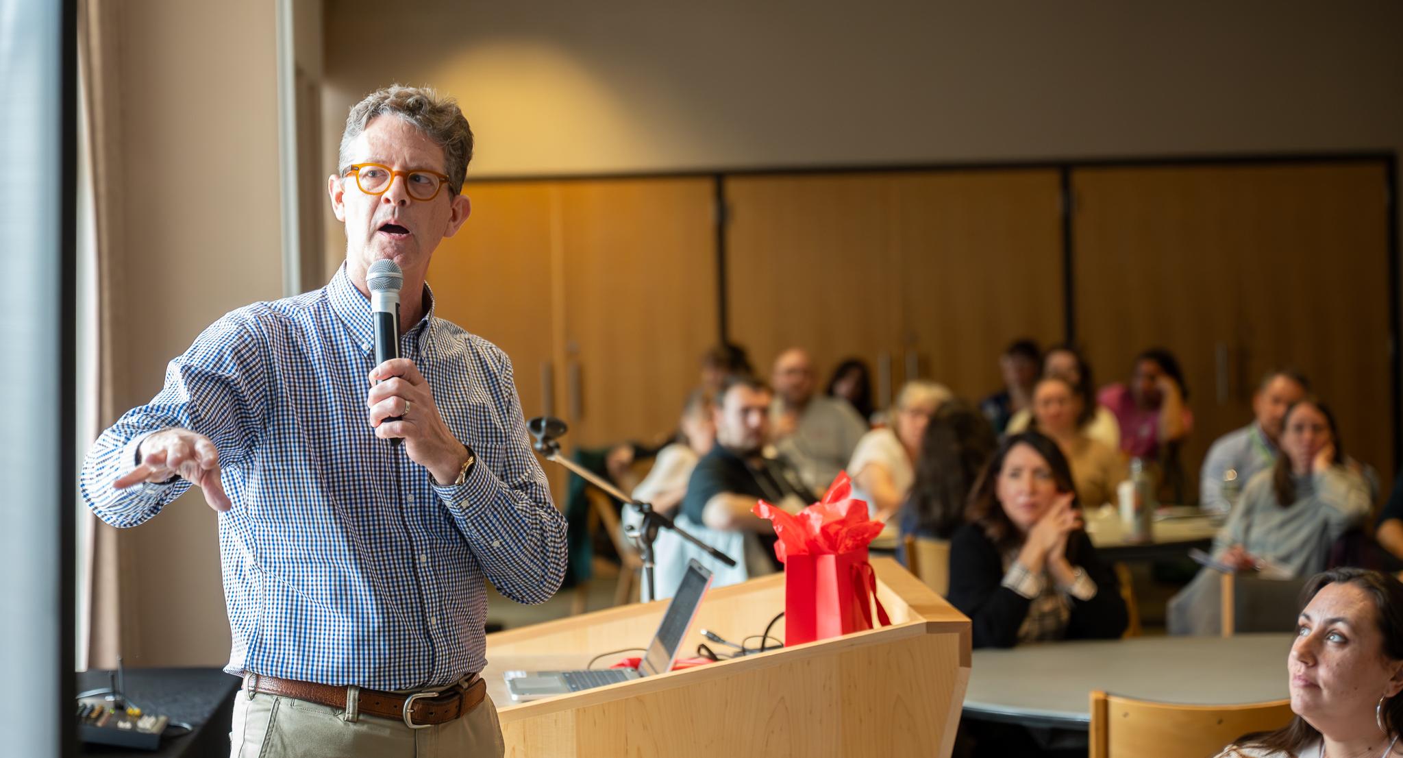 A man, holding a microphone, gives remarks in front of an audience in a classroom, while looking at a screen.