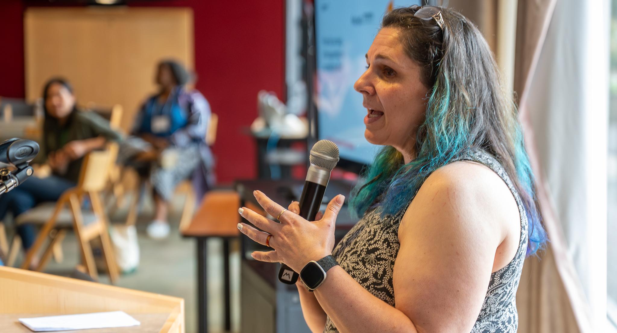 A person with dark hair, dyed blue at the end, gives remarks to a crowd in a classroom.