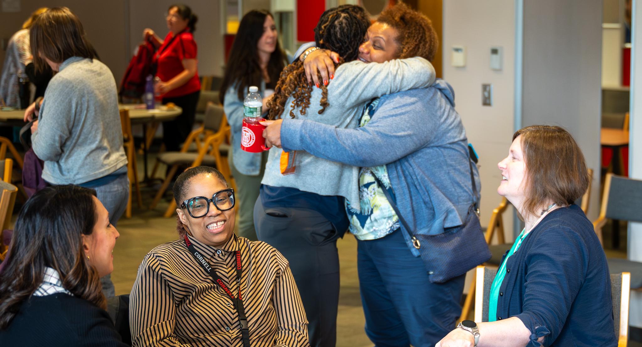 Two people, both wearing blue shirts and blue pants, hug next to a table of three people who are sitting at an event.