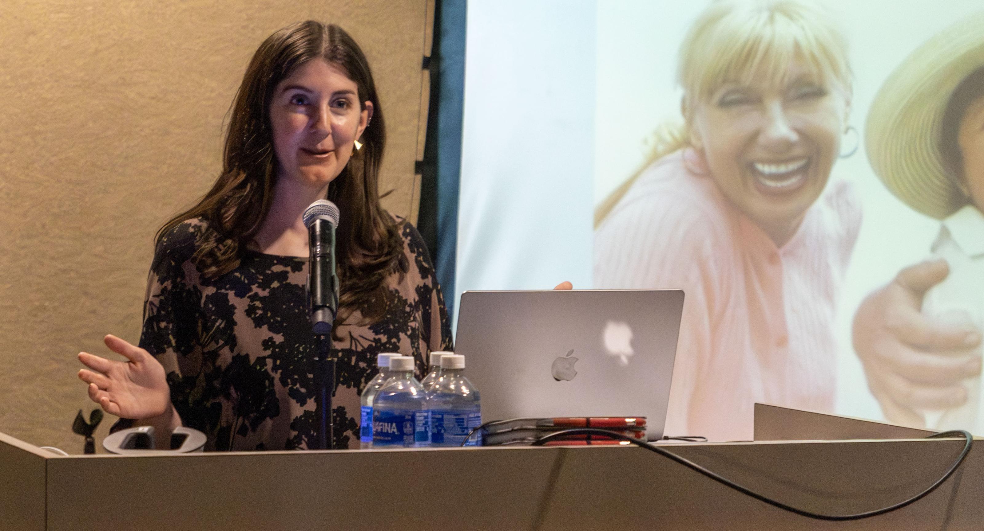 A woman makes a presentation at a dias, in front of a screen that is showing a smiling woman in one of her slides.