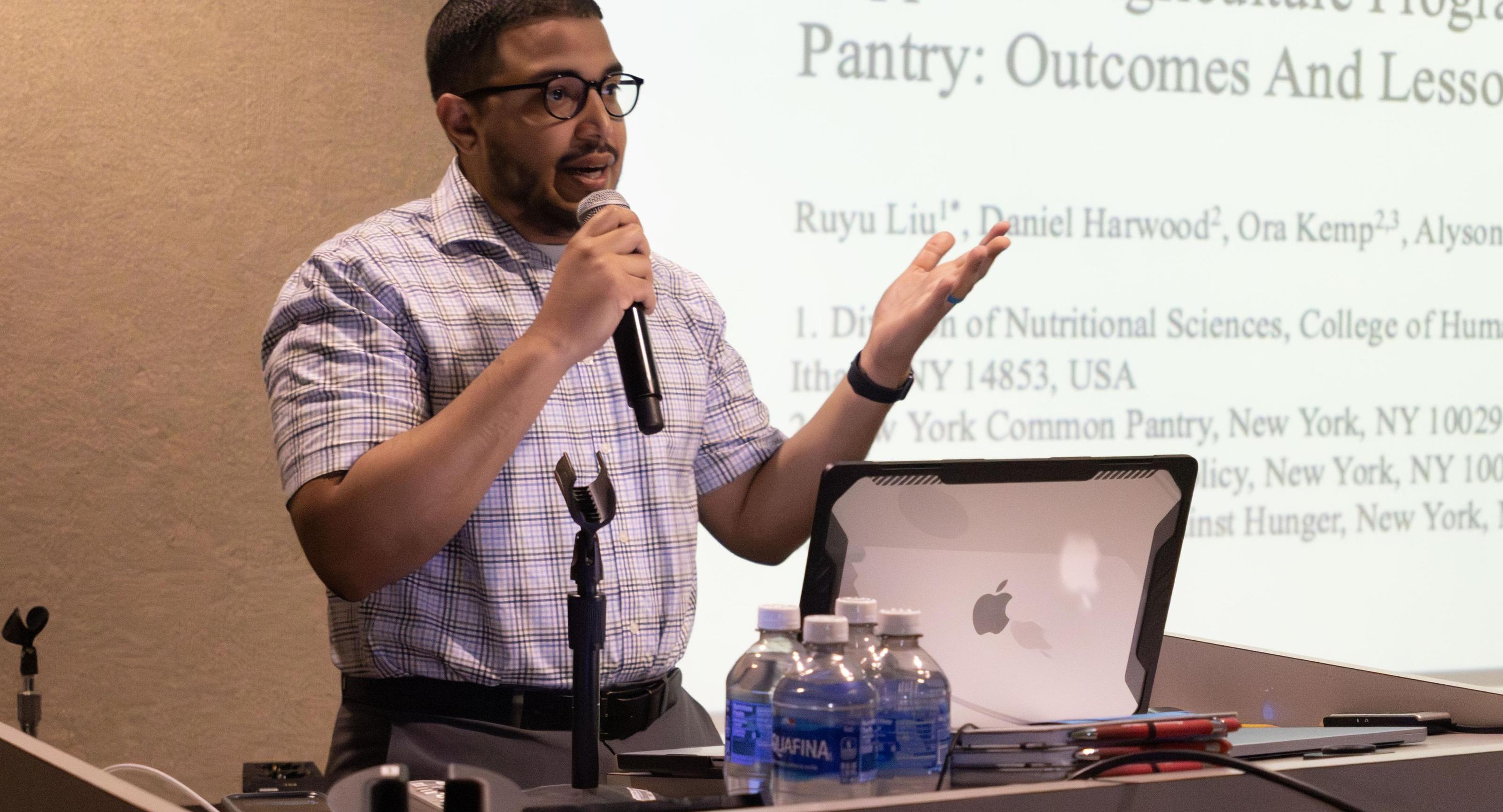 A man in pattenred shirt, holding a microphone and standing in front of a screen, gives a talk.