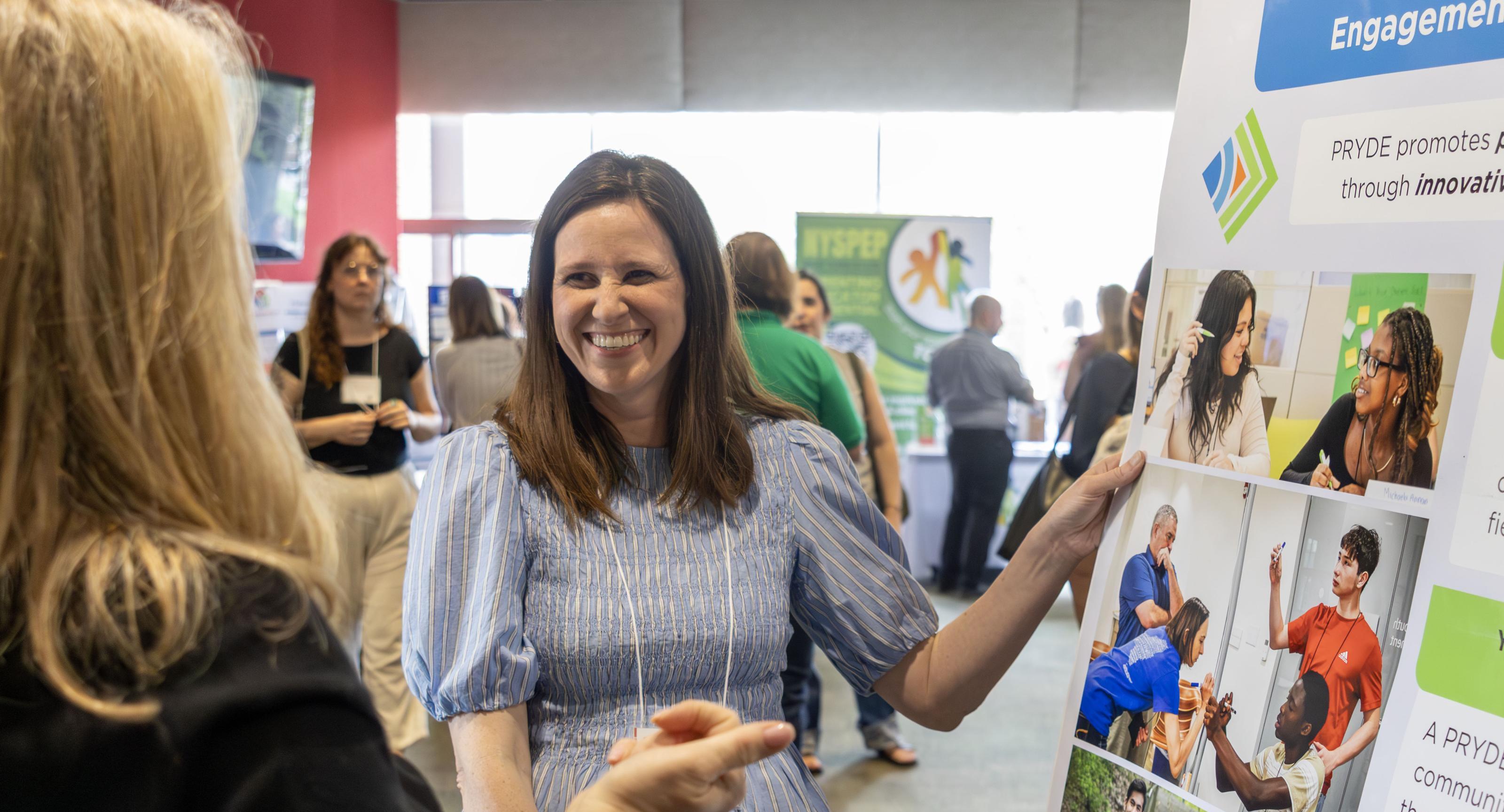 A woman wearing a blue and white blouse stands in front of her poster smiling at another person who is asking a quesiton.