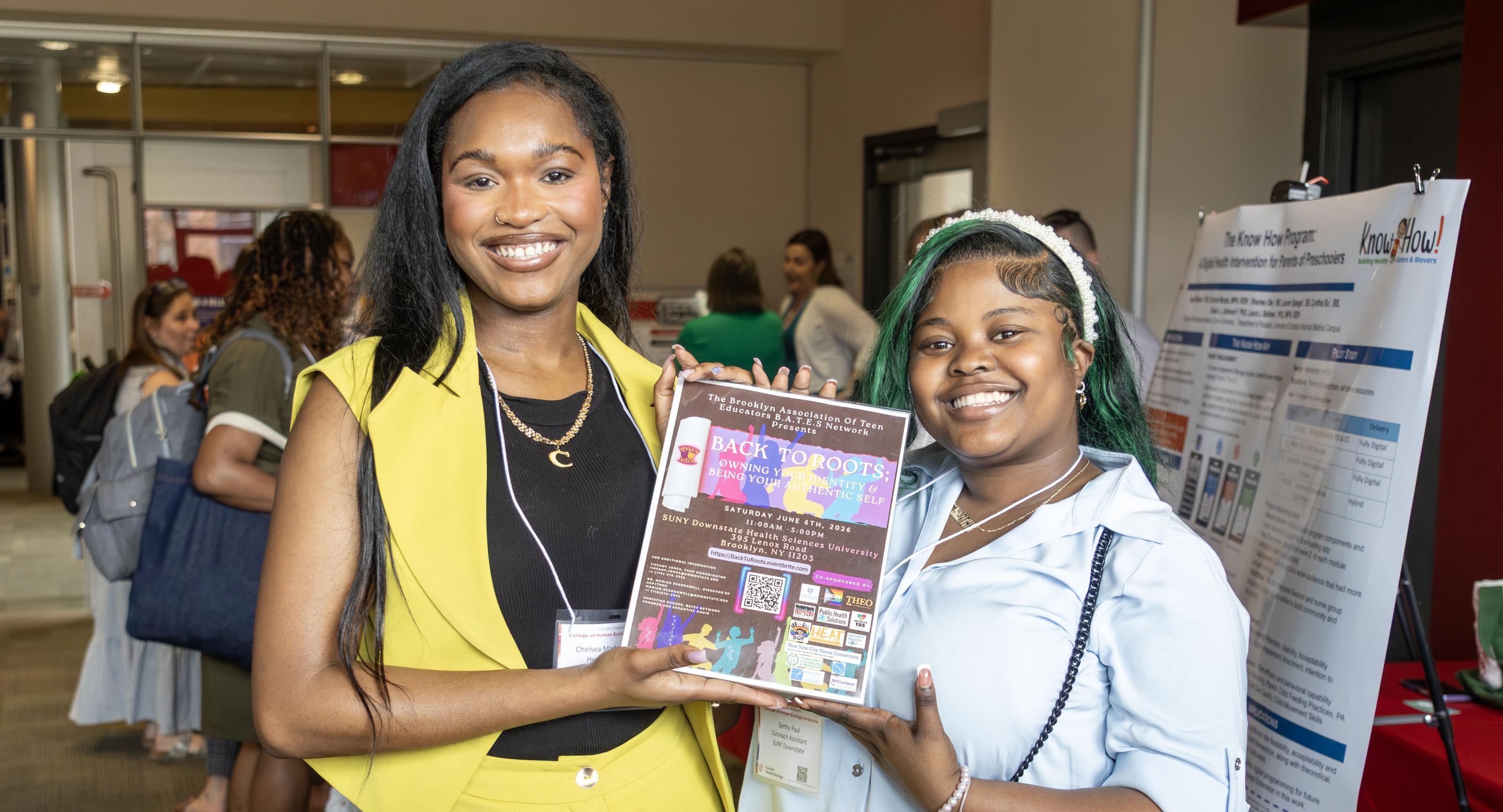 Two people stand next to each other holding up a small poster at an event. The person on the left is wearing a yellow vest with yellow pants and a black shirt, the person on the right is wearing a blue button down shirt with grey and white striped pants. 