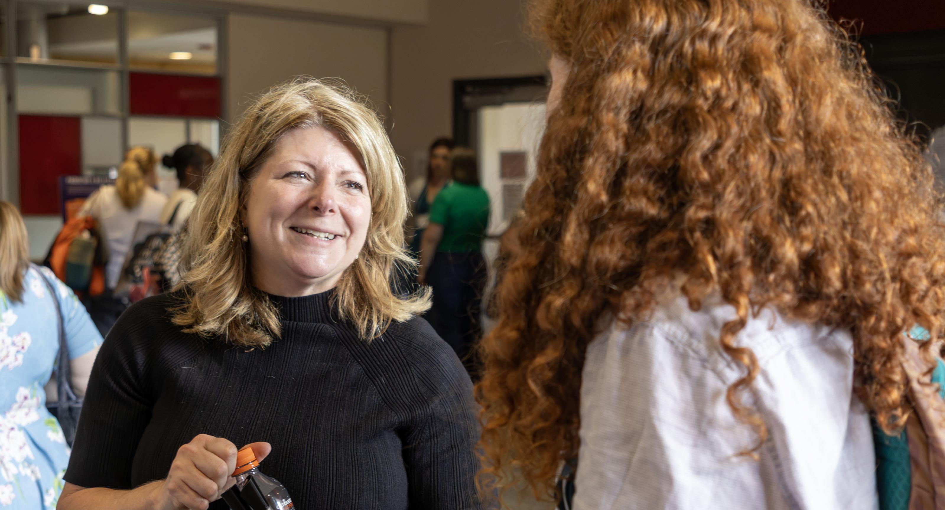 One woman, with blonde hair and a black shirt, holding a bottle, speaks with an unknown person with long curly red hair and a grey shirt.