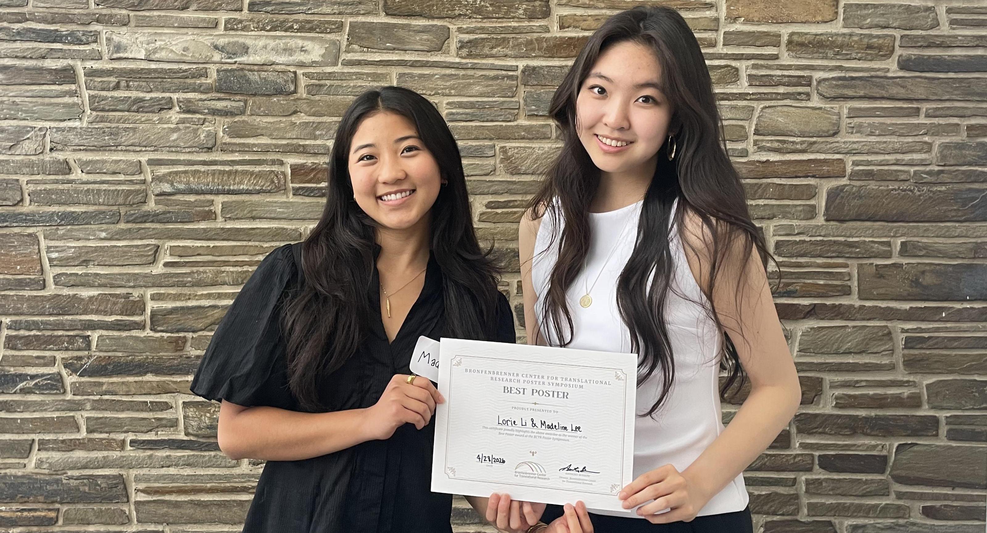 Two women stand in front of a stone facade holding a certificate.