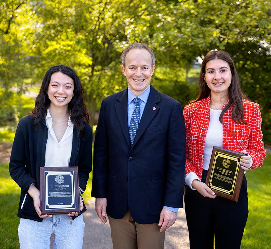 two students holding plaques with a man standing in the middle and trees behind them